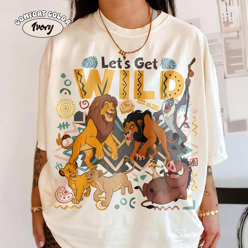 May include: Ivory Comfort Color t-shirt with a colorful graphic featuring characters from The Lion King and the text "Let's Get WILD". The design includes lions, a warthog, and a monkey, with decorative patterns.