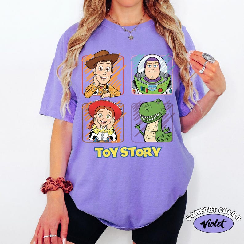 May include: A violet Comfort Colors t-shirt featuring a graphic of Toy Story characters: Woody, Buzz Lightyear, Jessie, and Rex. The text "TOY STORY" is printed below the characters. The shirt has short sleeves.