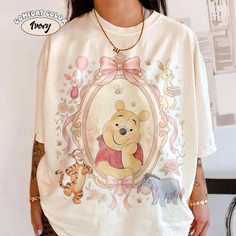 May include: Ivory-coloured t-shirt with a framed illustration of Winnie the Pooh and friends. The design includes Piglet with a balloon, Tigger, Eeyore, and Rabbit. The shirt has the words "Comfort Color" and "Ivory" printed on it.