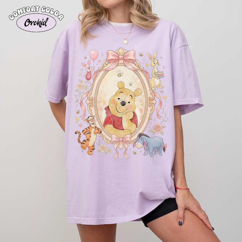May include: A lilac Comfort Color t-shirt with a framed illustration of Winnie the Pooh and friends. The design includes a pink bow, bees, and the text "Comfort Color Orchid".