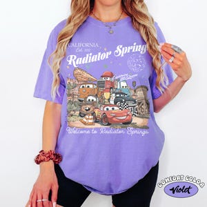K&ouml;nnte beinhalten: Ein violettes Comfort Colors T-Shirt mit einer Grafik von Charakteren aus dem Film Cars. Das Design enth&auml;lt den Text "California Est. 2012", "Radiator Springs" und "Welcome to Radiator Springs". Das Shirt hat einen Rundhalsausschnitt und kurze &Auml;rmel.