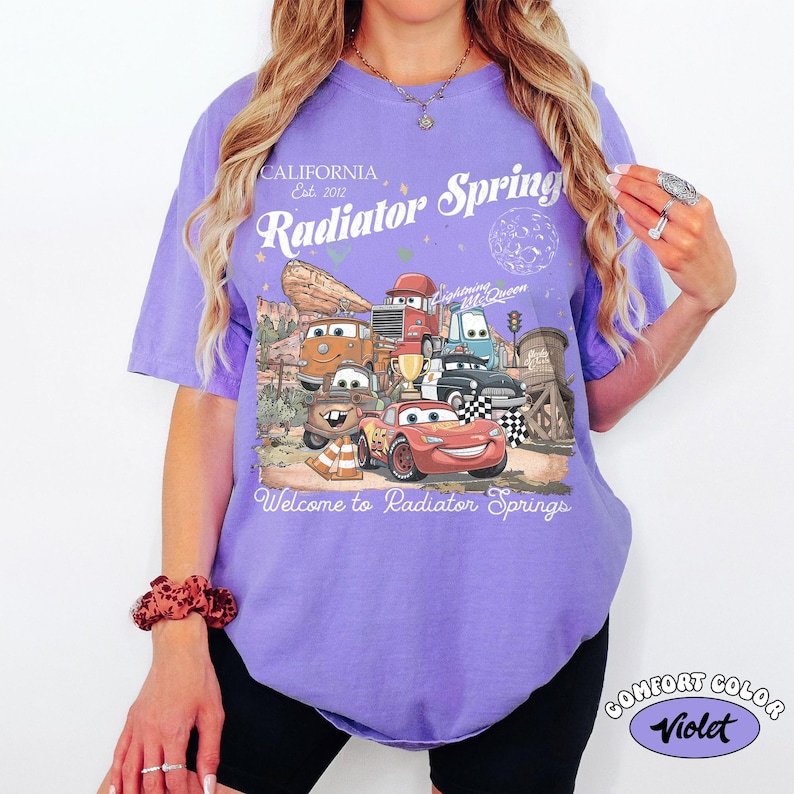 K&ouml;nnte beinhalten: Ein violettes Comfort Colors T-Shirt mit einer Grafik von Charakteren aus dem Film Cars. Das Design enth&auml;lt den Text "California Est. 2012", "Radiator Springs" und "Welcome to Radiator Springs". Das Shirt hat einen Rundhalsausschnitt und kurze &Auml;rmel.
