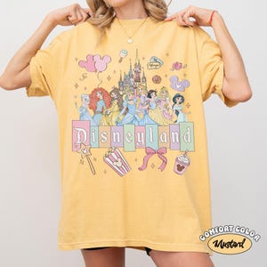 May include: Mustard yellow t-shirt featuring a Disneyland graphic with Disney princesses, a castle, and other themed icons. The text "Disneyland" is displayed in pastel colors. The shirt also has the words "Comfort Color" and "Mustard" printed on it.