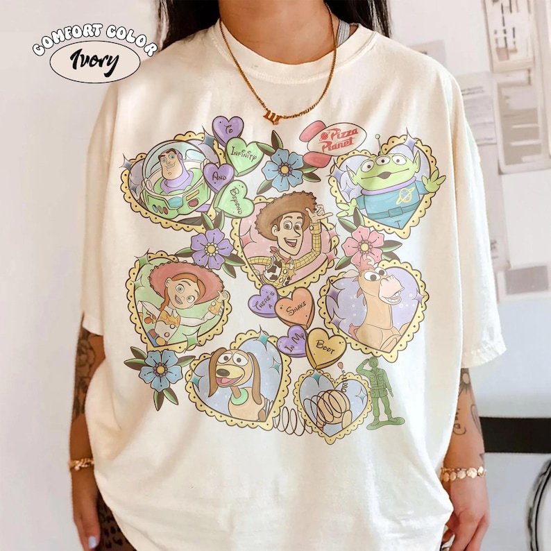May include: Ivory Comfort Color t-shirt featuring a colorful graphic of Toy Story characters in heart-shaped frames. The design includes Buzz Lightyear, Woody, Jessie, and others, with floral accents and the words "Pizza Planet".
