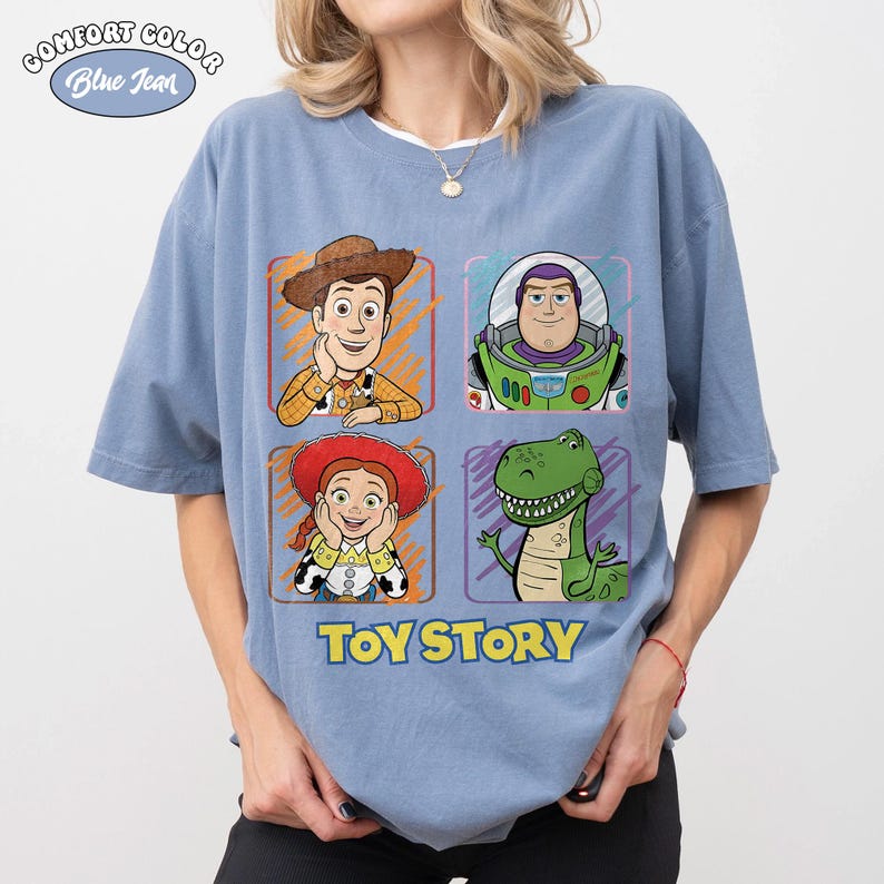 May include: Blue t-shirt featuring four cartoon character portraits from Toy Story: Woody, Buzz Lightyear, Jessie, and Rex. The words "TOY STORY" are printed in blue below the characters. The shirt has the words "COMFORT COLOR Blue Jean" printed on the top left.
