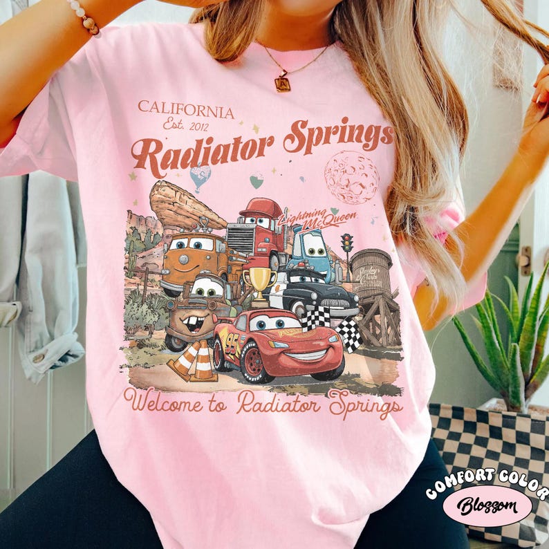 K&ouml;nnte beinhalten: Hellrosa Comfort Colors T-Shirt mit einer Grafik aus dem Film Cars, einschlie&szlig;lich Lightning McQueen. Das Design enth&auml;lt den Text "California Est. 2012" und "Radiator Springs". Das Shirt hat auch den Text "Welcome to Radiator Springs".