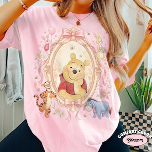 May include: Pale pink t-shirt with a framed illustration of Winnie the Pooh and friends, including Piglet, Tigger, Eeyore, and Rabbit. The design incorporates bows, flowers, and bees. The text "COMFORT COLOR Blossom" is printed on the bottom right.