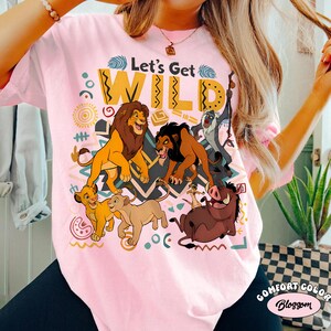 May include: Pink t-shirt featuring a graphic of characters from The Lion King, including Simba, Mufasa, Scar, Timon, Pumbaa, and Zazu. The text "Let's Get WILD" is printed above the characters in yellow and white.