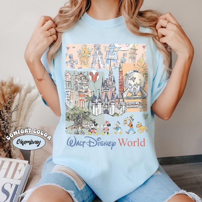 Peut inclure: T-shirt bleu clair avec un motif color&eacute; des monuments et personnages de Walt Disney World. Le motif comprend le ch&acirc;teau de Cendrillon, Mickey Mouse, Minnie Mouse, Donald Duck et Dingo. Les mots "Walt Disney World" sont imprim&eacute;s en dessous.