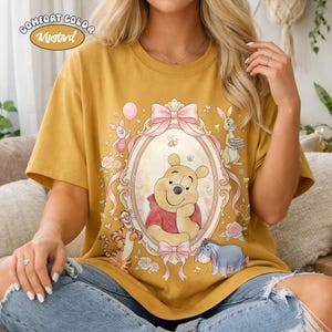 May include: Mustard-coloured t-shirt featuring a framed illustration of Winnie the Pooh with pink bows and surrounding characters. The shirt has the words "Comfort Color" and "Mustard" printed on it.