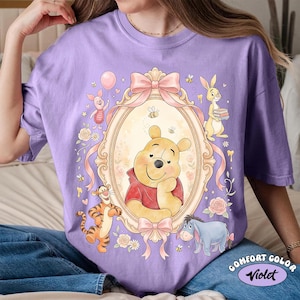 May include: A lilac Comfort Color t-shirt with a framed illustration of Winnie the Pooh and friends. The design includes Piglet, Tigger, Rabbit, and Eeyore, with bees, bows, and floral accents. The text "Comfort Color Violet" is at the bottom.