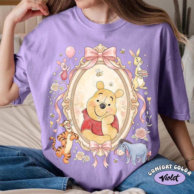 May include: A lilac Comfort Color t-shirt with a framed illustration of Winnie the Pooh and friends. The design includes Piglet, Tigger, Rabbit, and Eeyore, with bees, bows, and floral accents. The text "Comfort Color Violet" is at the bottom.