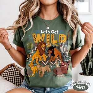 May include: Moss green t-shirt featuring a graphic of characters from The Lion King, including lions, a warthog, and a monkey. The text "Let's Get WILD" is displayed above the characters. The shirt is labeled "Comfort Color Moss".