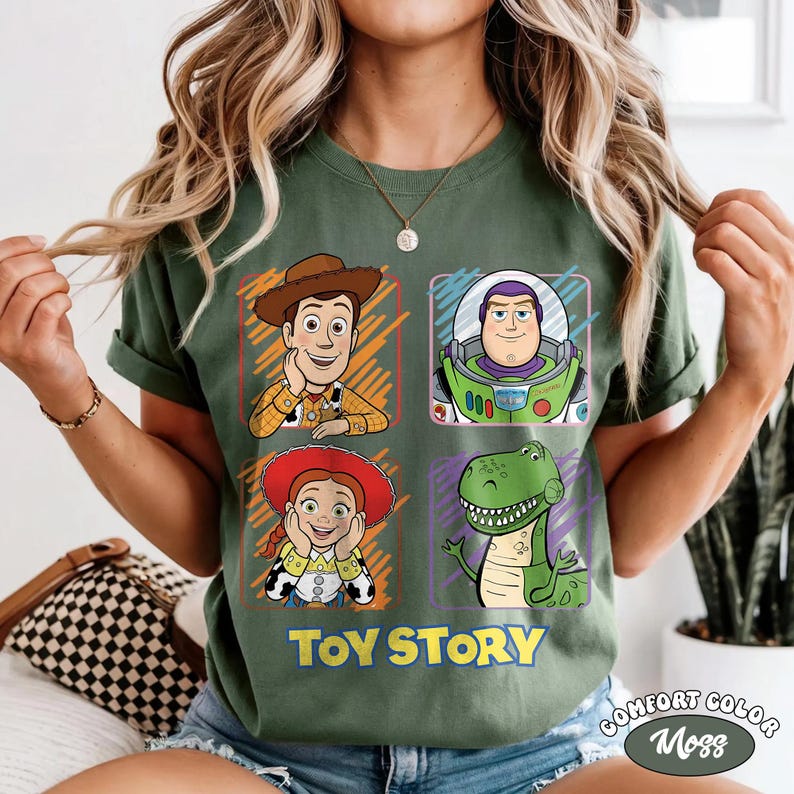 May include: Green t-shirt featuring four character portraits from the animated film "Toy Story": Woody, Buzz Lightyear, Jessie, and Rex. The words "Toy Story" are printed in yellow and blue. The shirt is a Comfort Colors Moss.