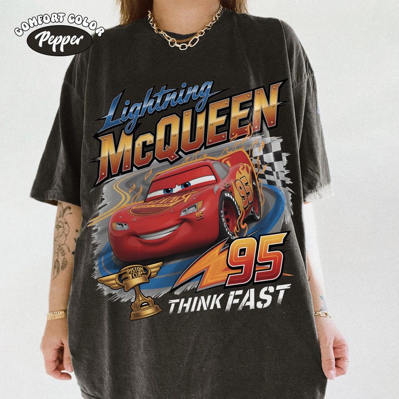Op de afbeelding: Donkergrijs t-shirt met Lightning McQueen uit de film Cars. Het ontwerp bevat de tekst "Lightning McQueen", "95" en "Think Fast", samen met een afbeelding van de rode raceauto en een geblokte vlag.