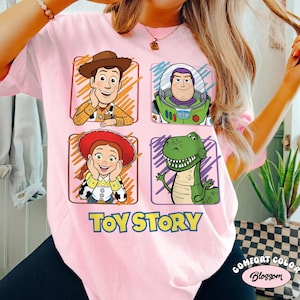 May include: A light pink t-shirt featuring four cartoon character portraits from the movie Toy Story: Woody, Buzz Lightyear, Jessie, and Rex. The words "Toy Story" are printed in yellow and blue below the characters.