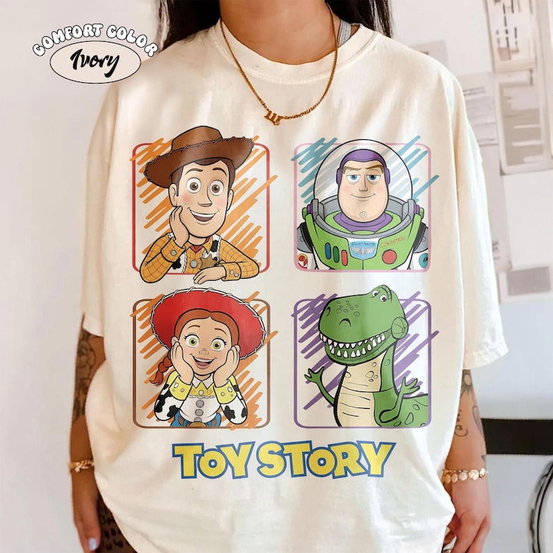 May include: Ivory Comfort Color t-shirt featuring four cartoon character portraits from Toy Story: Woody, Buzz Lightyear, Jessie, and Rex. The words "TOY STORY" are printed in blue below the characters. The shirt has a crew neck and short sleeves.