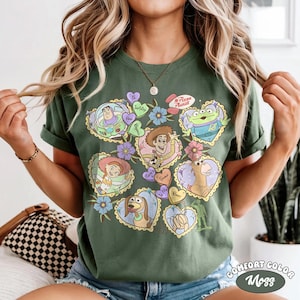 May include: Green Comfort Colors t-shirt featuring a colorful graphic of Toy Story characters in heart-shaped designs. The design includes Buzz Lightyear, Woody, and other characters, with floral accents and the text "Pizza Planet".