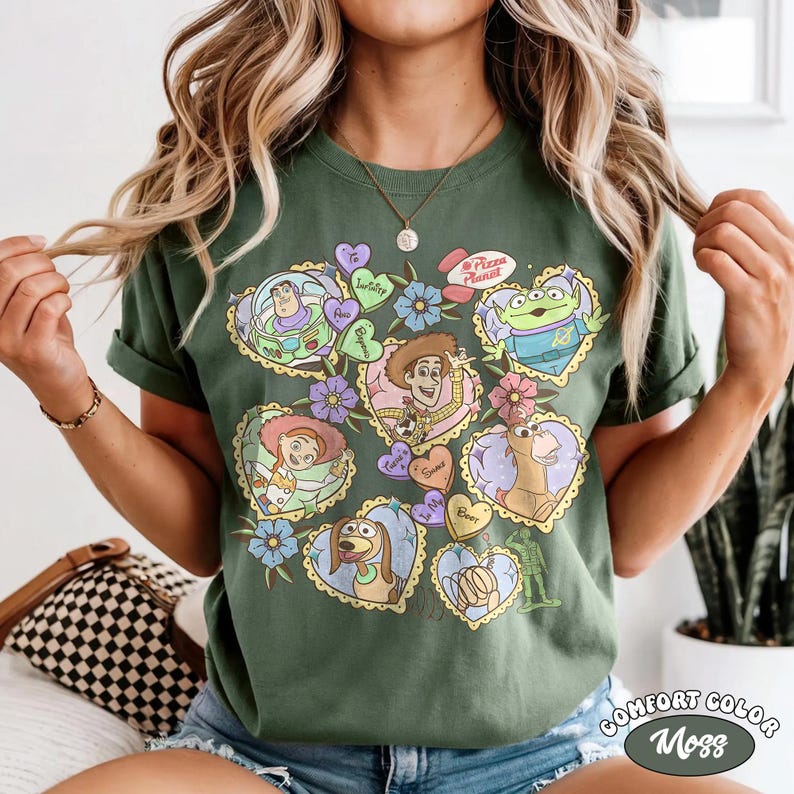May include: Green Comfort Colors t-shirt featuring a colorful graphic of Toy Story characters in heart-shaped designs. The design includes Buzz Lightyear, Woody, and other characters, with floral accents and the text "Pizza Planet".