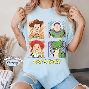 May include: Light blue t-shirt featuring four cartoon character portraits from the movie Toy Story: Woody, Buzz Lightyear, Jessie, and Rex. The words "Toy Story" are printed in yellow below the characters. The shirt is made of Comfort Color Chambray material.