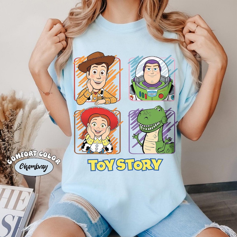 May include: Light blue t-shirt featuring four cartoon character portraits from the movie Toy Story: Woody, Buzz Lightyear, Jessie, and Rex. The words "Toy Story" are printed in yellow below the characters. The shirt is made of Comfort Color Chambray material.