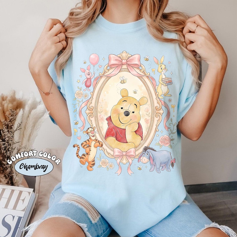 May include: Pale blue t-shirt with a framed illustration of Winnie the Pooh and friends. The design includes Pooh Bear, Piglet, Tigger, Eeyore, and Rabbit, surrounded by pink bows, flowers, and bees. The shirt has the words "Comfort Color Chambray".