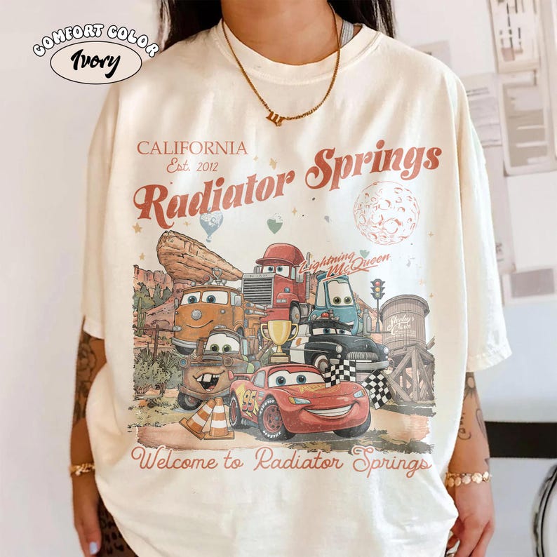 K&ouml;nnte beinhalten: Elfenbeinfarbenes T-Shirt mit einer Grafik mit Charakteren aus dem Film Cars. Das Design enth&auml;lt den Text "CALIFORNIA Est. 2012" und "Radiator Springs" im Vintage-Stil. Der Satz "Welcome to Radiator Springs" ist ebenfalls vorhanden.