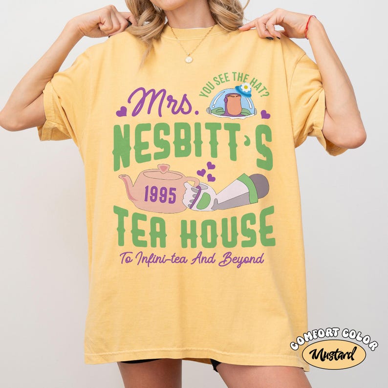 May include: Mustard-colored t-shirt with a graphic design featuring the text "Mrs. Nesbitt's Tea House" and a teapot illustration. The design includes the phrase "To Infini-tea And Beyond" and the year 1995.