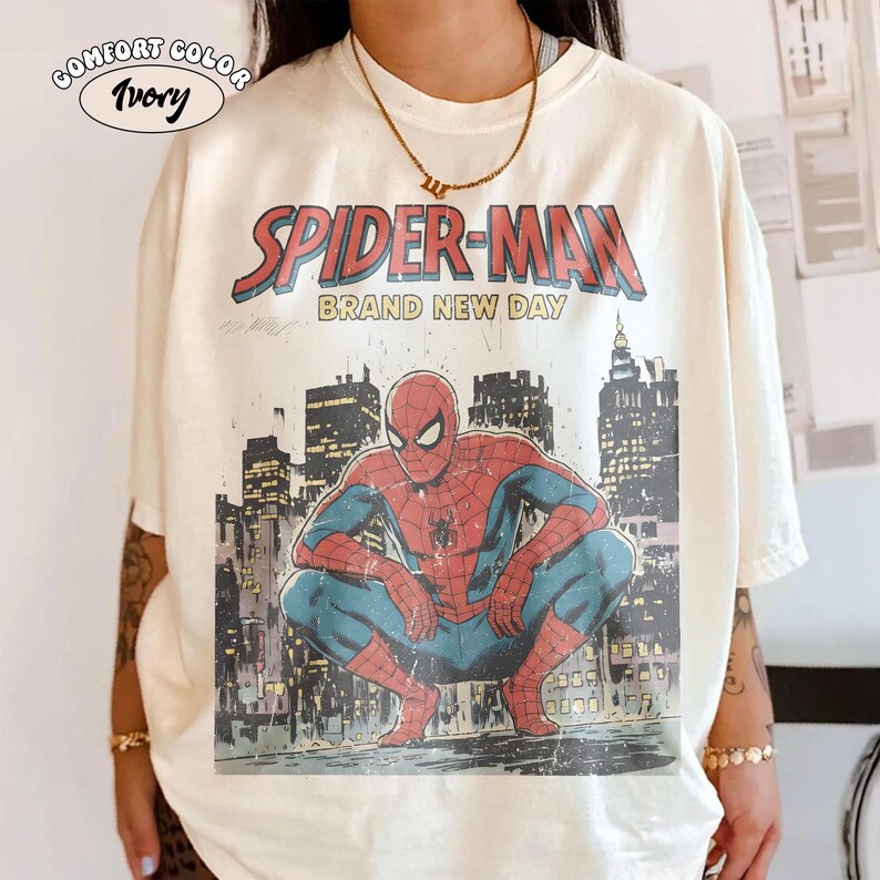 Puede incluir: Camiseta de color marfil con un gr&aacute;fico vintage de Spider-Man. El dise&ntilde;o incluye las palabras "SPIDER-MAN BRAND NEW DAY" sobre una ilustraci&oacute;n de Spider-Man agachado frente a un horizonte urbano. La camiseta tiene una etiqueta Comfort Color.
