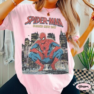 Puede incluir: Camiseta rosa claro con un dise&ntilde;o de Spider-Man agachado frente a un paisaje urbano. El texto "SPIDER-MAN BRAND NEW DAY" est&aacute; impreso encima de la imagen. Marca Comfort Colors.