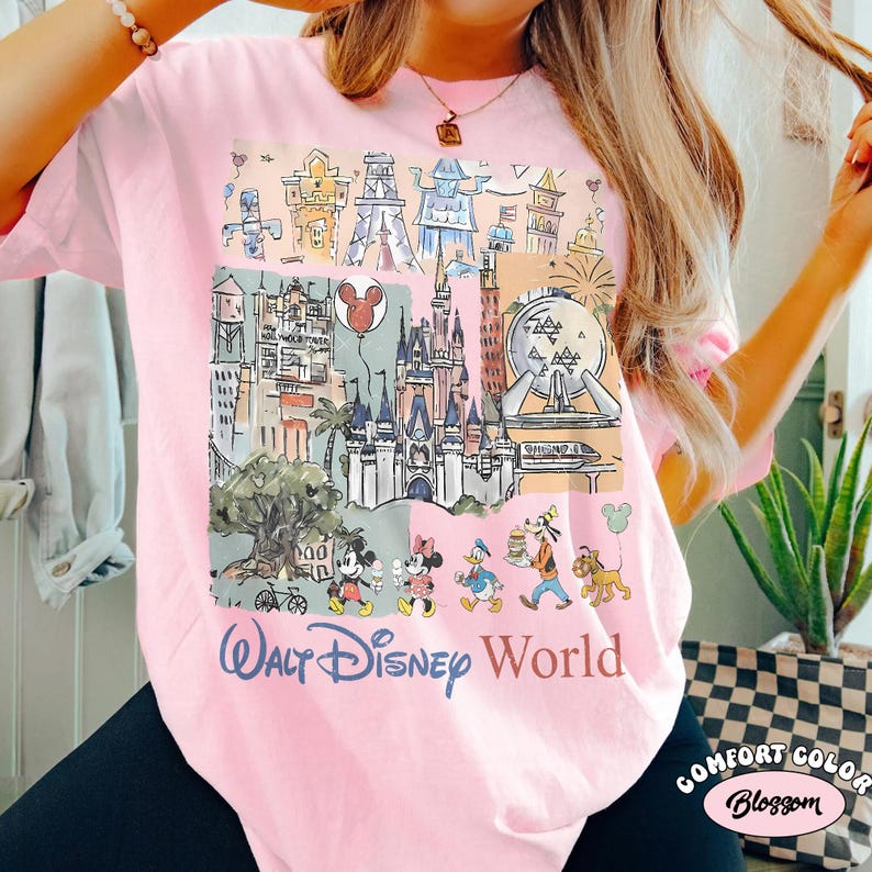 Peut inclure: T-shirt rose clair avec un motif color&eacute; des monuments et personnages de Walt Disney World, dont Mickey Mouse, Minnie Mouse et Donald Duck. Les mots "Walt Disney World" sont imprim&eacute;s en bleu. Le t-shirt est &eacute;tiquet&eacute; "Comfort Color Blossom".