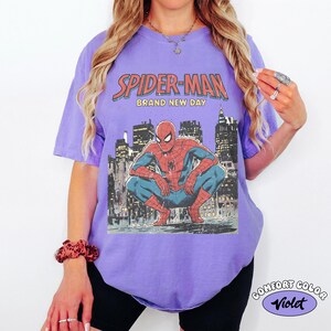 Puede incluir: Camiseta violeta con un gr&aacute;fico de Spider-Man agachado frente a un paisaje urbano. El texto "SPIDER-MAN BRAND NEW DAY" est&aacute; impreso arriba. La camiseta est&aacute; etiquetada como "Comfort Color Violet".