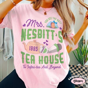 May include: A light pink t-shirt with the text "Mrs. Nesbitt's Tea House" in green and purple. The design includes a teapot, the year 1995, and a cartoon image of a character. The phrase "To Infini-tea And Beyond" is also included.