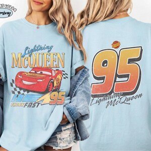Puede incluir: Camiseta azul claro con Lightning McQueen de la pel&iacute;cula Cars. El frente muestra el personaje, el texto "Lightning McQueen" y "95". La parte trasera presenta un gran "95" y el texto "Lightning McQueen".