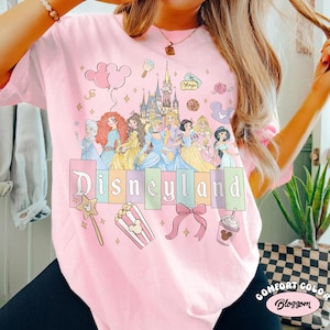 May include: Pink t-shirt with a Disneyland graphic featuring Disney princesses, a castle, and various icons. The word "Disneyland" is in pastel colors. Additional graphics include a wand, popcorn, a bow, and a drink.