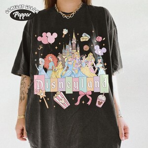 May include: Black t-shirt with a Disneyland graphic. The design features Disney princesses, a castle, and the word "Disneyland" in pastel colors. Additional graphics include Mickey Mouse balloons, popcorn, and a bow.