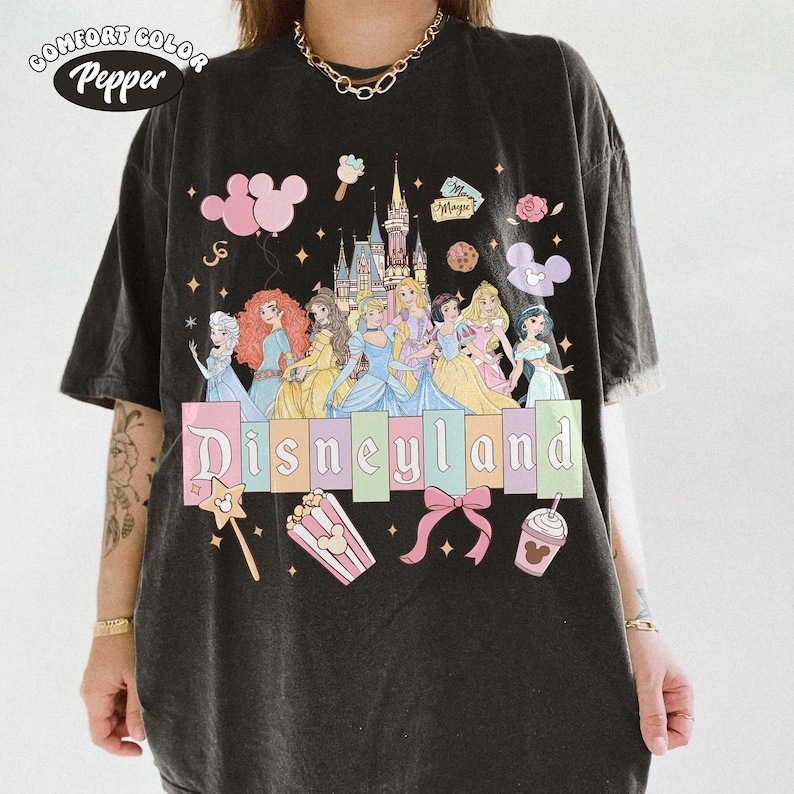 May include: Black t-shirt with a Disneyland graphic. The design features Disney princesses, a castle, and the word "Disneyland" in pastel colors. Additional graphics include Mickey Mouse balloons, popcorn, and a bow.