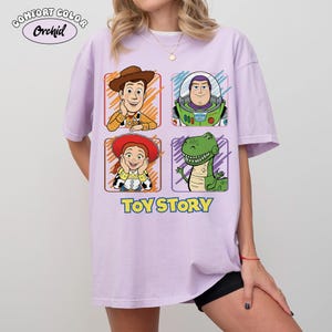 May include: A light purple t-shirt featuring four cartoon character portraits from Toy Story: Woody, Buzz Lightyear, Jessie, and Rex. The words "TOY STORY" are printed in yellow below the characters. The shirt also has the text "COMFORT COLOR Orchid" at the top.
