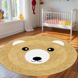 Pode incluir: Um tapete redondo de juta bege claro, com um desenho de rosto de urso. O rosto do urso apresenta um focinho branco, olhos pretos e um nariz preto. O tapete está colocado sobre um chão de madeira num quarto infantil.
