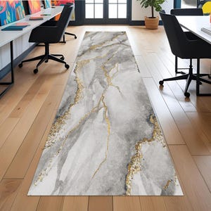 Può includere: Un lungo tappeto rettangolare con un design simile al marmo. Il tappeto presenta una miscela di toni grigi e bianchi, con venature color oro. Il tappeto è posizionato su un pavimento in legno in un ambiente ufficio.