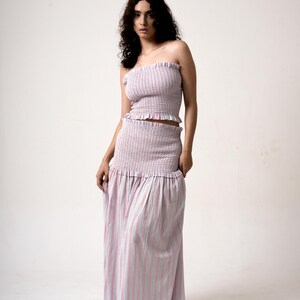 May include: A two-piece outfit featuring a strapless top and a floor-length skirt. The set is made of a fabric with vertical stripes in pink and light blue. The top has a ruffled edge, and the skirt has a gathered waist.