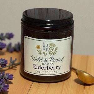 May include: A jar of elderberry-infused honey with a black lid and a label that reads "Wild & Rooted Goods." The label features floral illustrations. A gold spoon rests beside the jar, with lavender sprigs in the background.