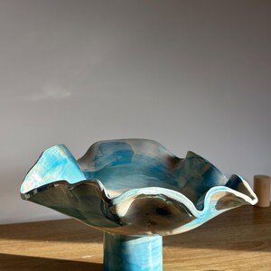Può includere: Una ciotola in ceramica su piedistallo con un bordo ampio e ondulato e una base cilindrica. La superficie è decorata con una pennellata astratta dipinta a mano in tonalità vivaci di turchese, azzurro e beige tenue, creando un effetto fluido.