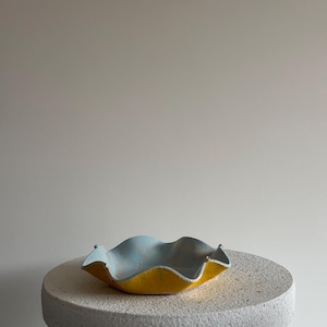 Op de afbeelding: Een handgemaakte keramische kom met een golvende rand. De kom heeft een felgele buitenkant en een lichtblauwe binnenkant. Hij staat op een gespikkelde, ronde betonnen sokkel. De achtergrond is effen neutraal.