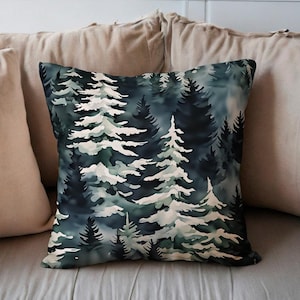 May include: Square decorative pillow featuring a watercolor forest scene in shades of blue, green, and white. The design depicts stylized evergreen trees, creating a serene, nature-inspired aesthetic. The pillow is placed on a bed with neutral-colored pillows.
