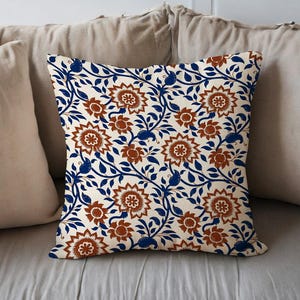 May include: Square decorative pillow with a floral pattern. The pillow features a cream-colored background with a repeating design of brown flowers and blue vines. The pillow is placed on a bed with other pillows.