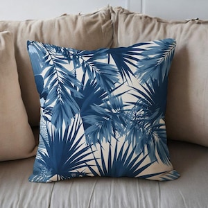 Puede incluir: Cojín cuadrado con un diseño de hojas tropicales azules y blancas. El cojín tiene un fondo beige y está rodeado de otros cojines. El patrón presenta varios tonos de azul, creando un efecto de capas.