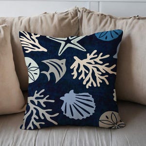 Puede incluir: Cojín decorativo cuadrado con fondo azul oscuro y un patrón de vida marina blanca, azul claro y beige, incluyendo estrellas de mar, coral y conchas. El cojín está colocado sobre una superficie beige claro.