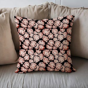 Puede incluir: Cojín cuadrado con fondo negro y un patrón repetitivo de diseños florales blancos y rojos. El cojín está colocado sobre una cama con almohadas de colores neutros y una colcha gris claro.