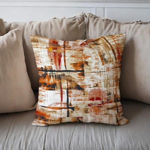 Op de afbeelding: Vierkant sierkussen met een abstract ontwerp in tinten oranje, bruin, zwart en wit. Het kussen heeft penseelstreken en ligt op een neutraal getint bed met andere kussens.